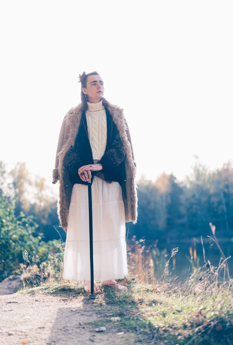 Aramis stützt sich auf einen Gehstock. Er hebt sich in Mantel und Kleid vom Himmel ab.