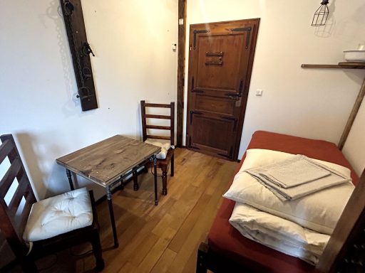 Kleines Zimmer mit einem Tisch, zwei Stühlen und einem Bett. Holzfußboden und Tür.