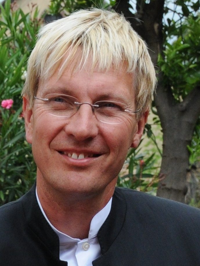 Porträt eines lächelnden Mannes mit blondem Haar und Brille, im Freien vor einer Pflanze.