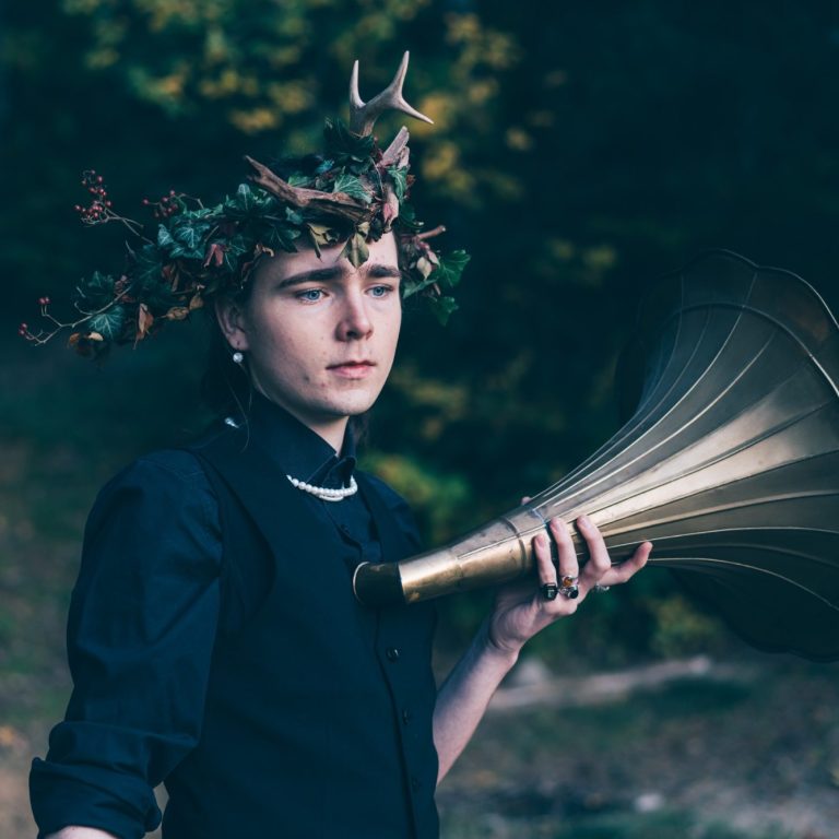 Aramis Freiherr von Maltzahn hält sich einen Grammophonlautsprecher an die Brust. Er trägt eine Krone aus Zweigen und schwarze Kleider.