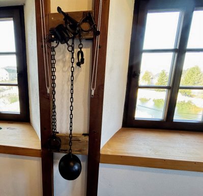 Historischer Kran mit Kette und Gewicht an einer Wand mit Fenstern.