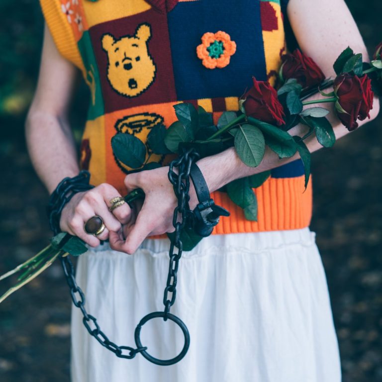 Nahaufnahme eines Oberkörpers in kindlicher Kleidung. Viele Ringe an den Fingern umklammern dornige Rosen und sind mit ihnen verkettet.