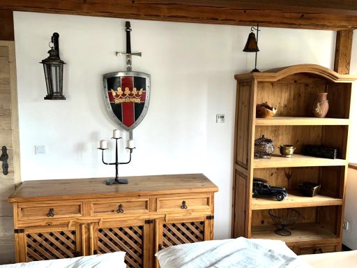 Holzkommode mit Kerzenständer, Wand mit Schild und Schwert, Regal mit Keramik.